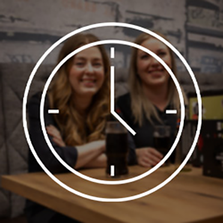 Zwei Frauen sitzen entspannt mit Getränken an einem Tisch und lachenin die Kamera. Überlagert wird das Bild von einem weißes Piktogram einer Uhr. 