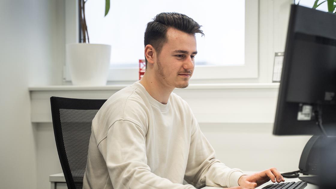 Ein junger Mitarbeiter mit dunklen Haaren arbeitet konzentriert an einem PC-Arbeitsplatz im Büro. Im Hintergrund steht eine große Pflanze.