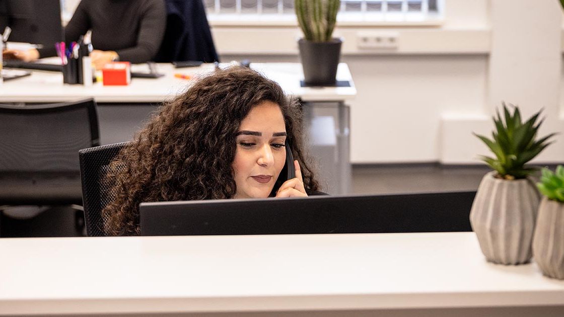 Eine weibliche Mitarbeiterin sitzt an einem Empfangsschalter im Büro und telefoniert. Sie hat lockige dunkle Haare und blickt konzentriert auf ihren Bildschirm. Im Hintergrund sind weitere Arbeitsplätze mit Computern und Pflanzen zu sehen.