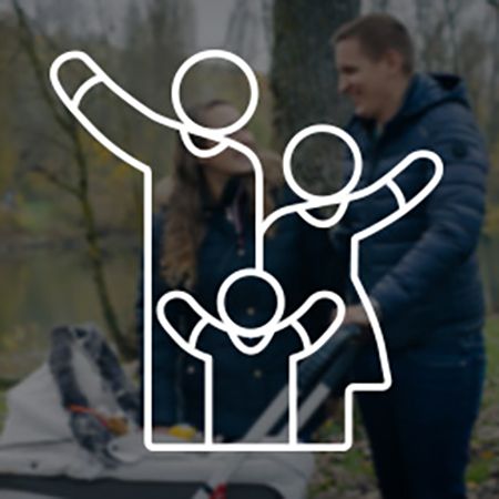 Eine junge Familie spaziert durch einen Park mit einem Kinderwagen. Im Hintergrund sind Bäume und ein See sichtbar. Die Eltern, beide in warmer Kleidung, lächeln und interagieren miteinander. Überlagert wird das Bild von einem weißen Piktogramm, das eine Familie mit einem Kind darstellt, bei dem alle Personen fröhlich die Arme heben.