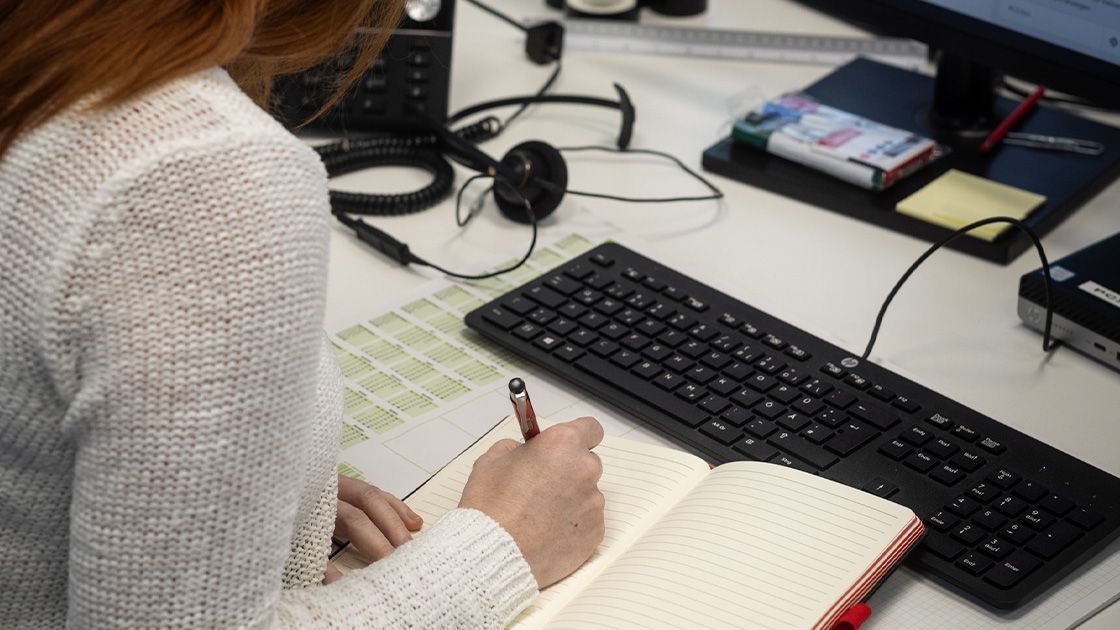 Eine Frau in einem weißen Strickpullover wird von hinten gezeigt, während sie in ein aufgeschlagenes Notizbuch schreibt. Auf dem Schreibtisch befinden sich eine Tastatur, ein Monitor, ein Telefon sowie Büromaterialien wie Haftnotizen und Stifte. Ein Headset liegt neben dem Telefon.